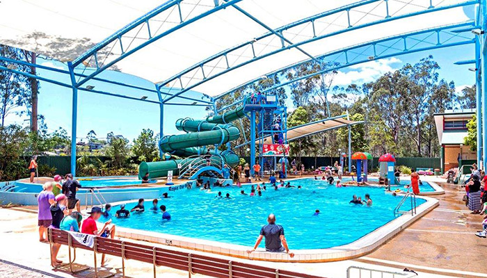 Level concrete concourse around a pool under a blue frame and white shade sail. Features a green spiral waterslide, wooden-slat bench seating, and stainless steel handrails for water entry.