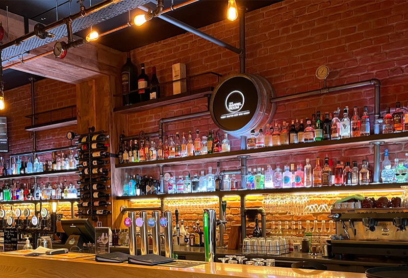 Level timber bar counter with silver beer taps. Backlit spirit shelves on a red brick wall with industrial pipe accents. Warm overhead lighting from hanging filament bulbs.