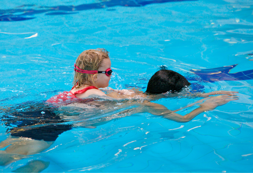 Child with blonde hair and red goggles on the back of a swimmer in a bright blue pool. Tiled floor and dark lane markings visible beneath the water's surface.