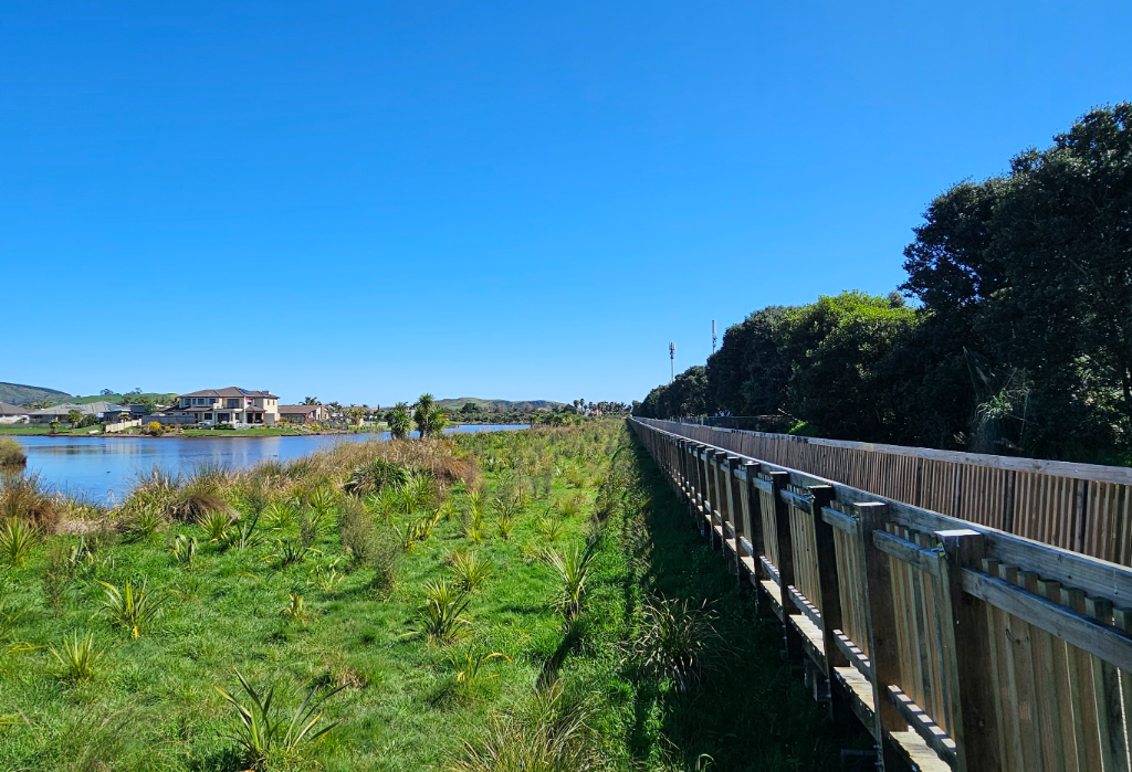 Long, level timber boardwalk with vertical slat handrails on both sides. The pathway consists of wooden planks and runs straight through a grassy field next to a lake.