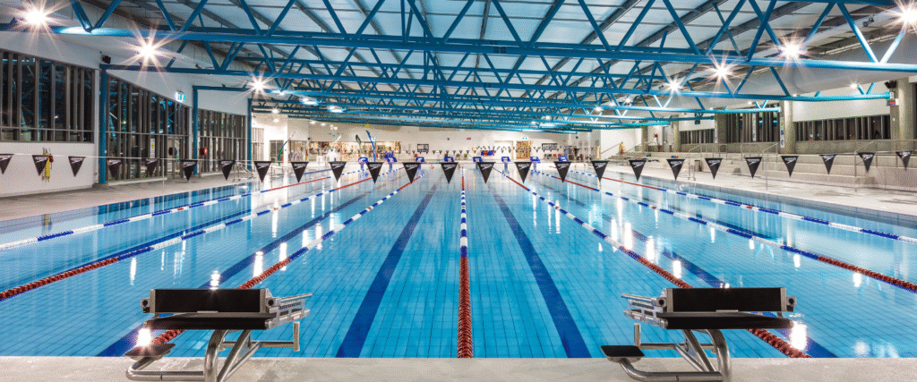 Large indoor pool with level deck and black starting blocks. Blue lanes marked with red and white ropes under bright overhead spotlights, a blue metal truss ceiling, and glass side walls.