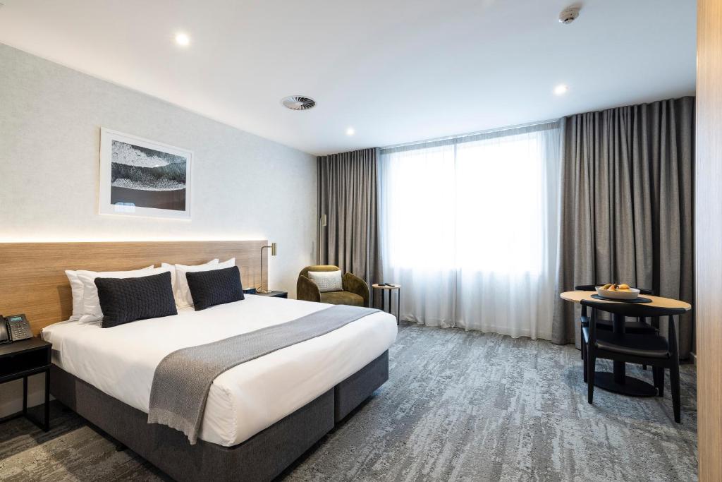 Level grey patterned carpet throughout. Large bed with clear floor space on three sides. Green armchair and a small dining table with two chairs. Well-lit with ceiling and natural light.