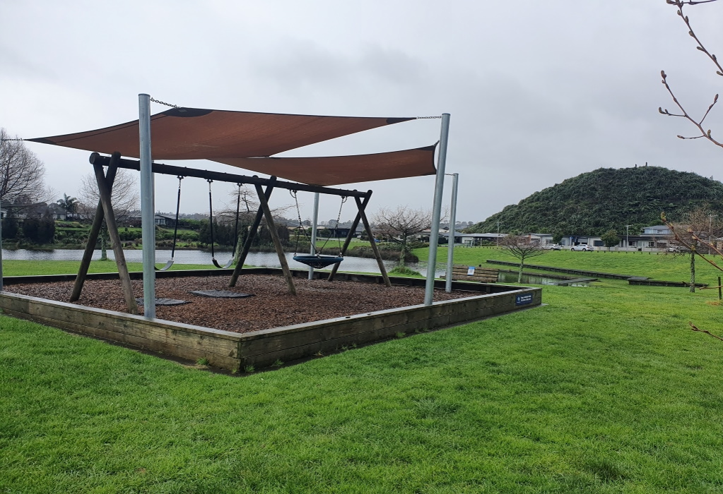 Timber-edged woodchip pit with two standard and one nest swing under a brown shade sail. No ramp access. Surrounded by level grass. A wooden park bench and small blue sign are visible nearby.