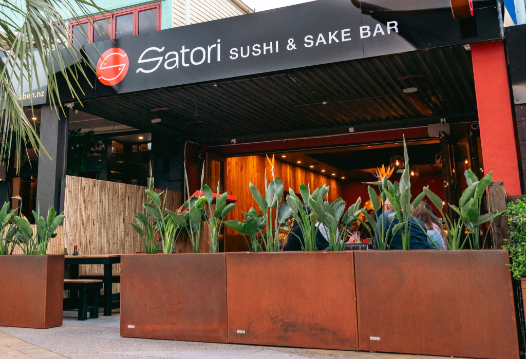 Level paved walkway leads to a wide, step-free entrance of Satori Sushi & Sake Bar. Inside, wooden tables and bench seating are on a flat ground surface. Large planters line the front boundary.