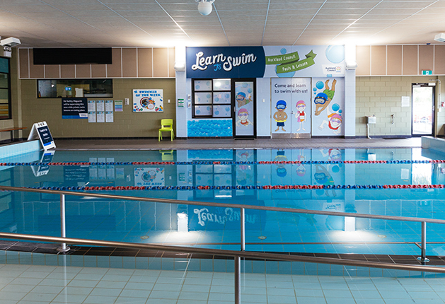 Silver handrails line a ramp into a blue indoor pool. A level tiled deck features a green chair and colourful 'Learn to Swim' wall signage under bright overhead lighting.