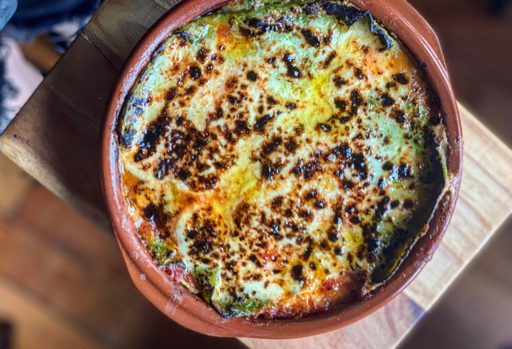 Round terracotta dish containing a baked meal. A bubbly white surface is heavily browned and charred in patches. Green vegetables and red sauce are visible around the edges.