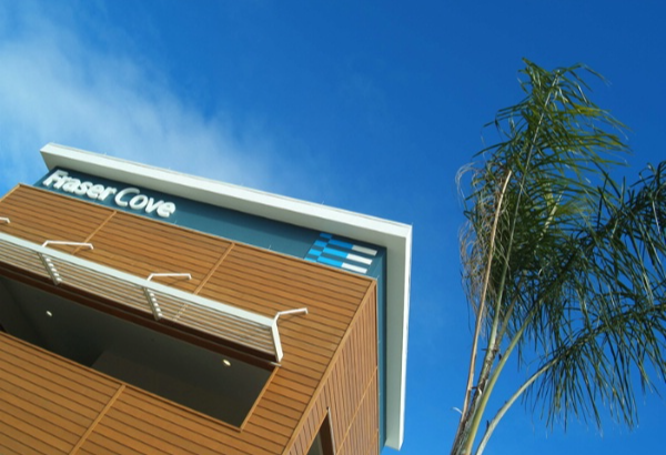 "Fraser Cove" sign in white lettering on a blue panel above brown timber-look cladding and a white metal sun louvre. A palm tree stands beside the building against a bright blue sky.
