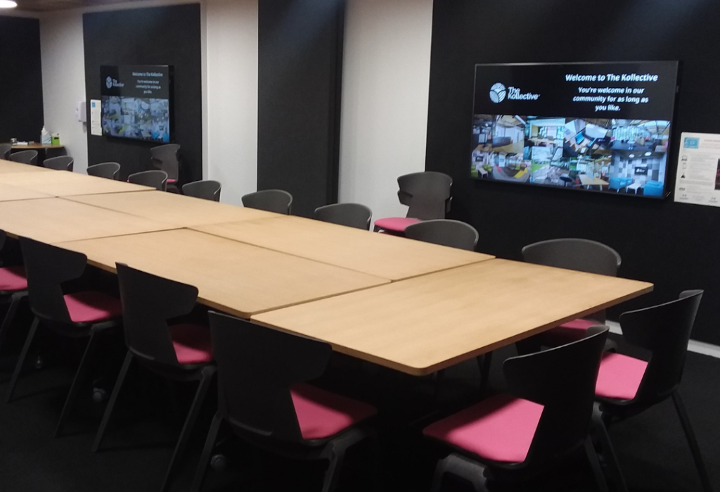 Long timber table with black-framed chairs and pink cushions on level dark carpet. Two wall-mounted screens. Open floor plan with wide pathways for movement around seating.
