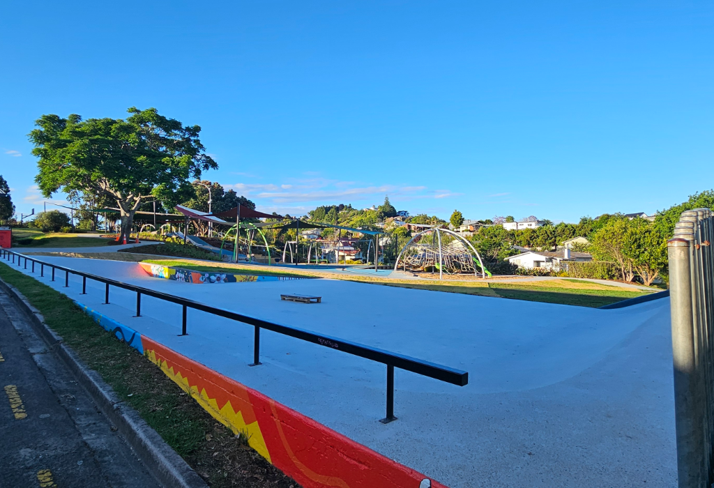 Smooth concrete skate park with a long black handrail and colourful red, yellow and orange painted border. Level paths connect to a playground with climbing equipment, swings and shade sails.