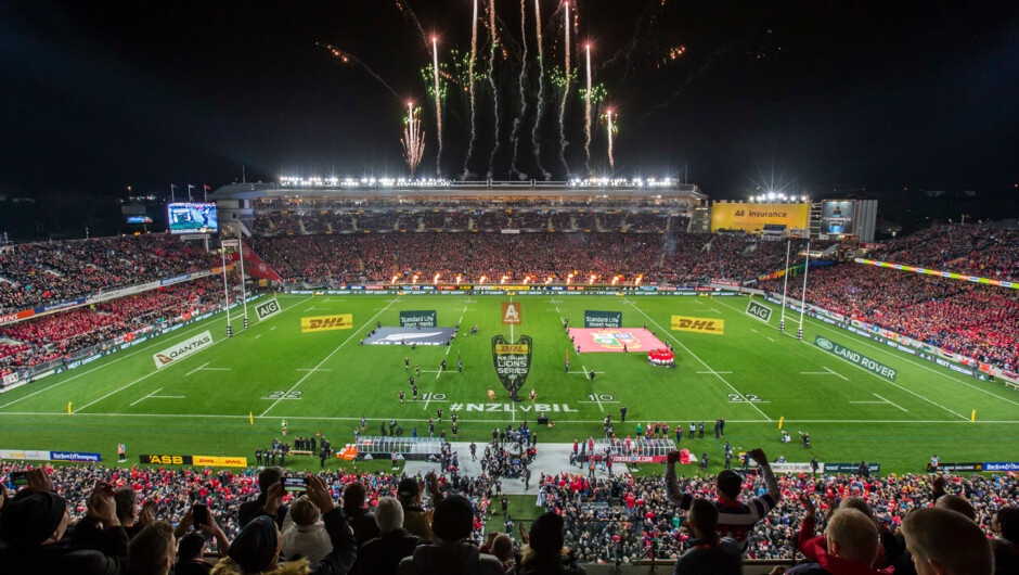 Tiered seating surrounds a green grass pitch with white markings and tall goal posts. Floodlights, screens, and signage illuminate the night venue with fireworks and crowds in the stands.
