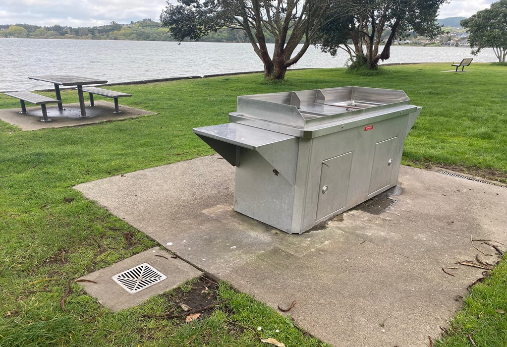 Stainless steel barbecue on a level concrete pad. Nearby metal picnic table with benches on a concrete base. Surrounding area is flat mown grass with a metal drain grate set into the ground.