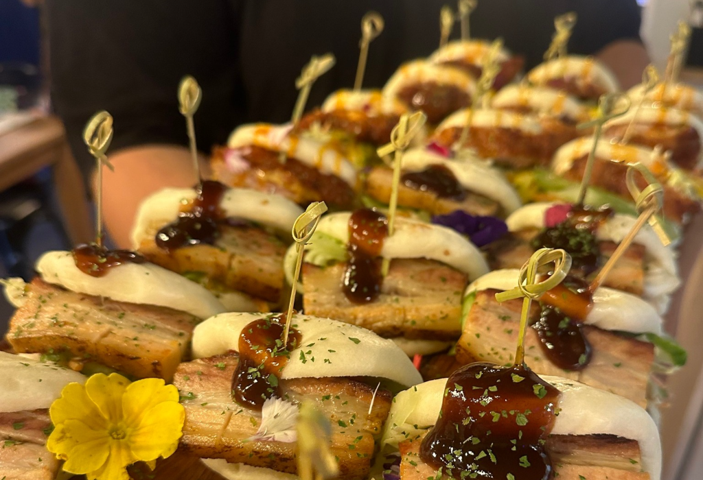 Bao buns filled with thick pork belly, topped with dark sauce and herbs. Each bun is held by a twisted bamboo skewer. Yellow and purple flowers decorate the platter.