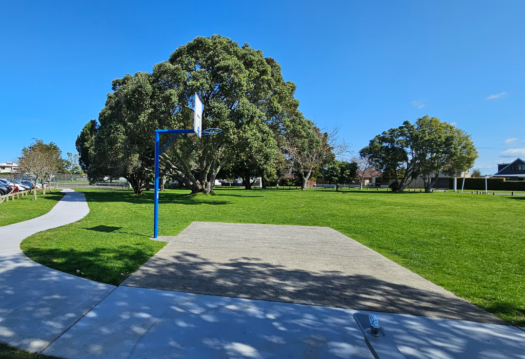 Level concrete pathway curves through a grassy park past a flat concrete basketball pad with a blue pole. A low-set metal drinking fountain is located beside the path.