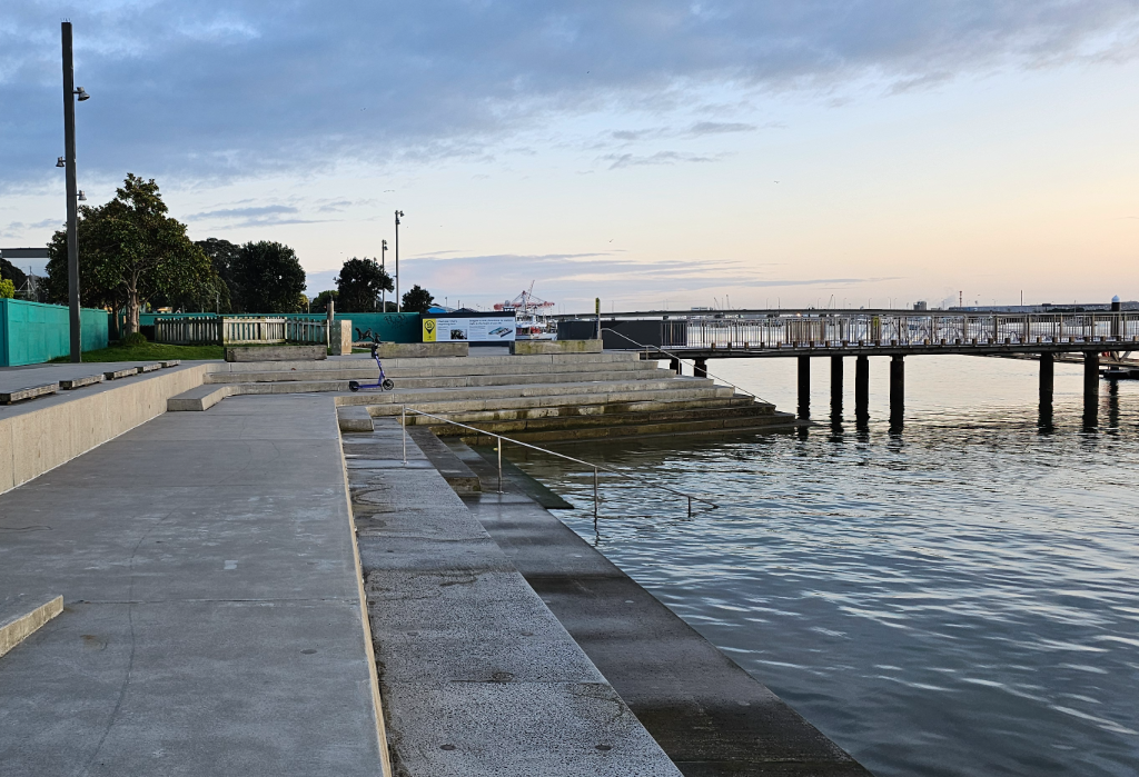 Wide, level concrete pathway beside tiered seating and a ramp with metal handrails leading into the water. A timber pier on pylons extends over the harbour. Grey concrete surfaces throughout.