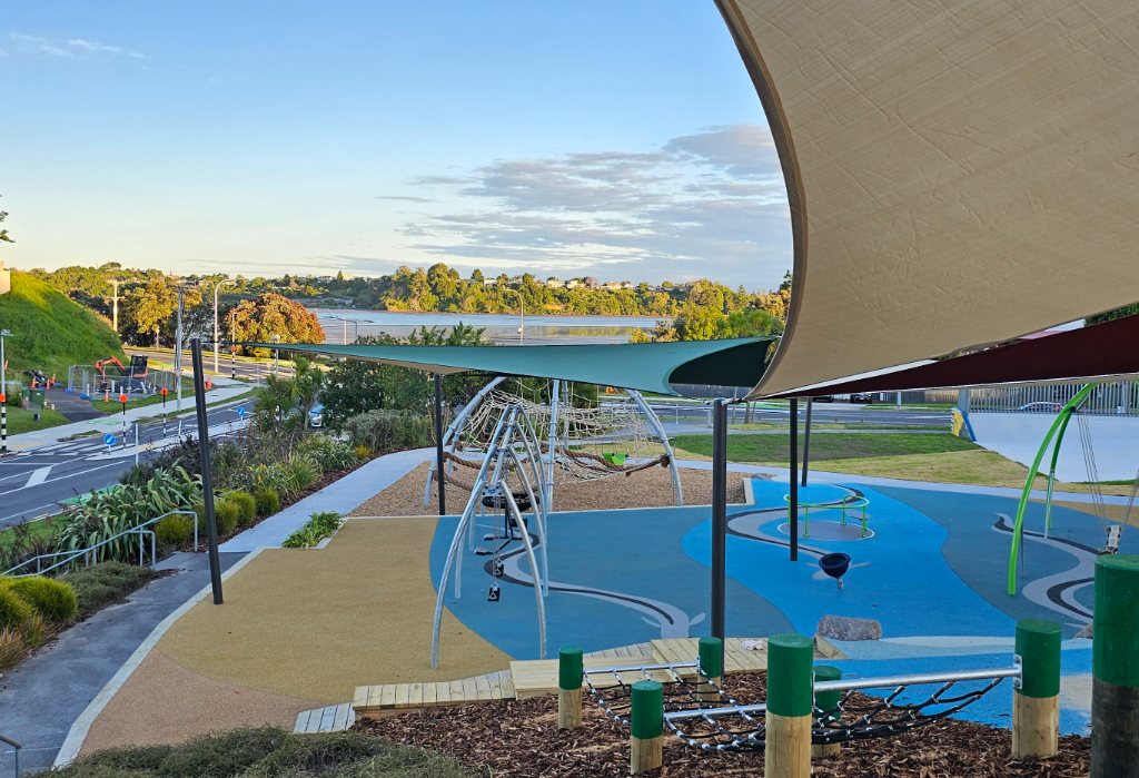 Concrete path with silver handrail leads to blue and yellow rubber soft-fall playground. Features shade sails, climbing nets, and swings with wood chip and grass borders overlooking water.