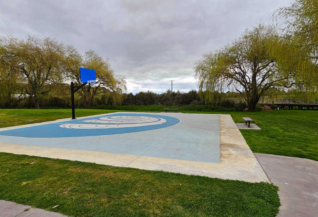 Level concrete pathway leads to a flat blue and grey basketball court with spiral markings. Two backless timber benches sit on level concrete pads adjacent to the court area.