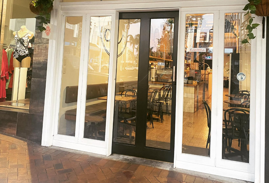 Level entrance with wide glass and black sliding doors from a red brick paver footpath. Inside features light-coloured flooring, wooden tables, chairs, and dark padded bench seating.