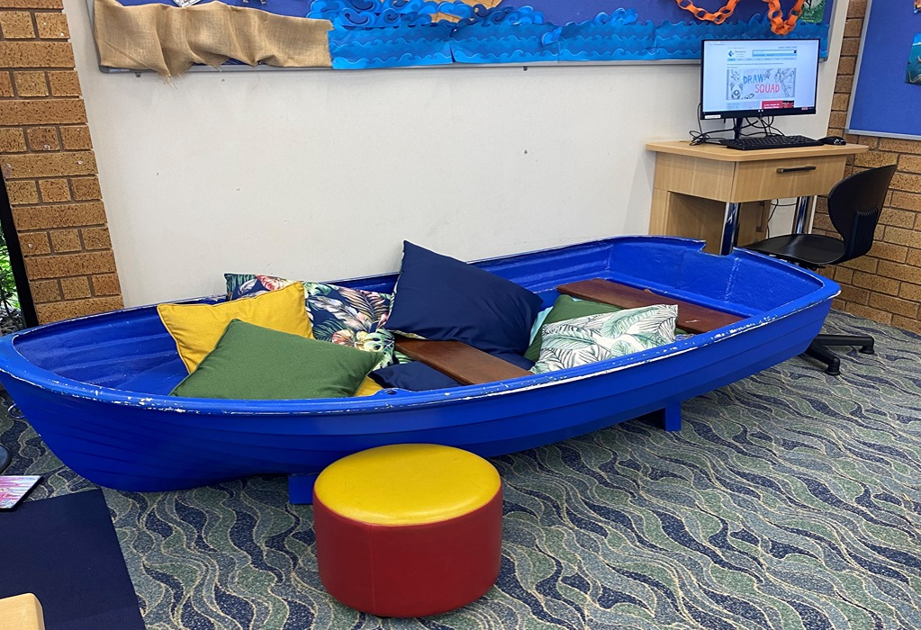 Blue rowboat seating with colourful cushions on a wavy blue and green patterned carpet. A red and yellow round ottoman is in front. Timber desk and black chair are in the background.