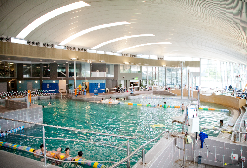 Indoor aquatic centre with tiled ramps and silver handrails for pool entry. High white arched ceiling with skylights. Turquoise pool with multi-coloured lane ropes and tiered seating on the right.
