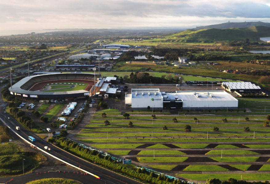 Level bitumen parking area beside a white arena. Wide paved roads and flat concrete pathways lead to building entrances. Large grassed area features dark, level dirt vehicle tracks.
