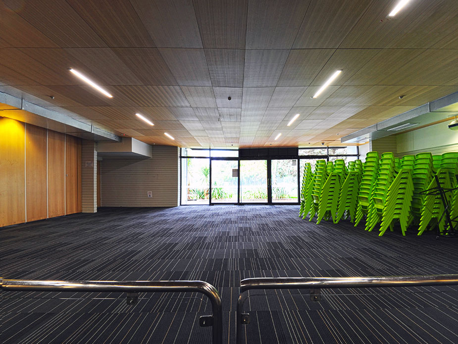 Large hall with level, dark grey striped carpet. Metal handrails in foreground lead toward glass double doors. Stacks of lime green chairs against right wall. Linear ceiling lighting.