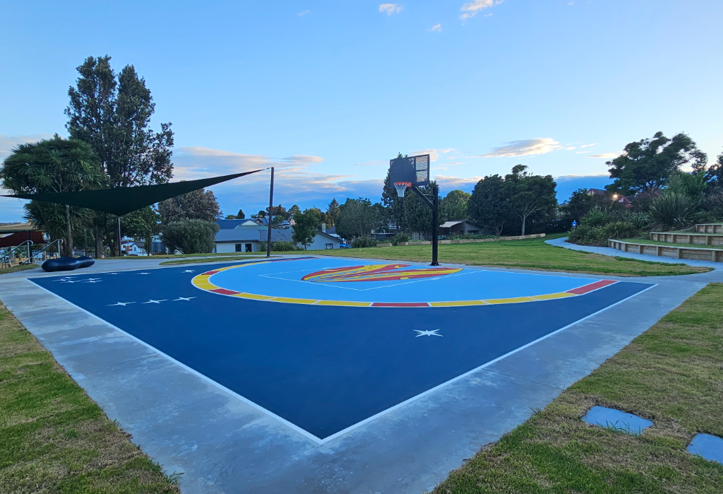 Level dark blue basketball court with yellow and red markings. A wide concrete path leads to tiered timber seating. A large shade sail covers a level area with a low-profile blue bench.