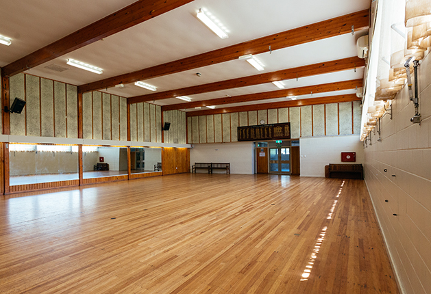 Large hall with level, polished timber floors and exposed ceiling beams. Long mirrors with timber handrails line one wall. Timber benches sit against masonry walls near a double-door exit.