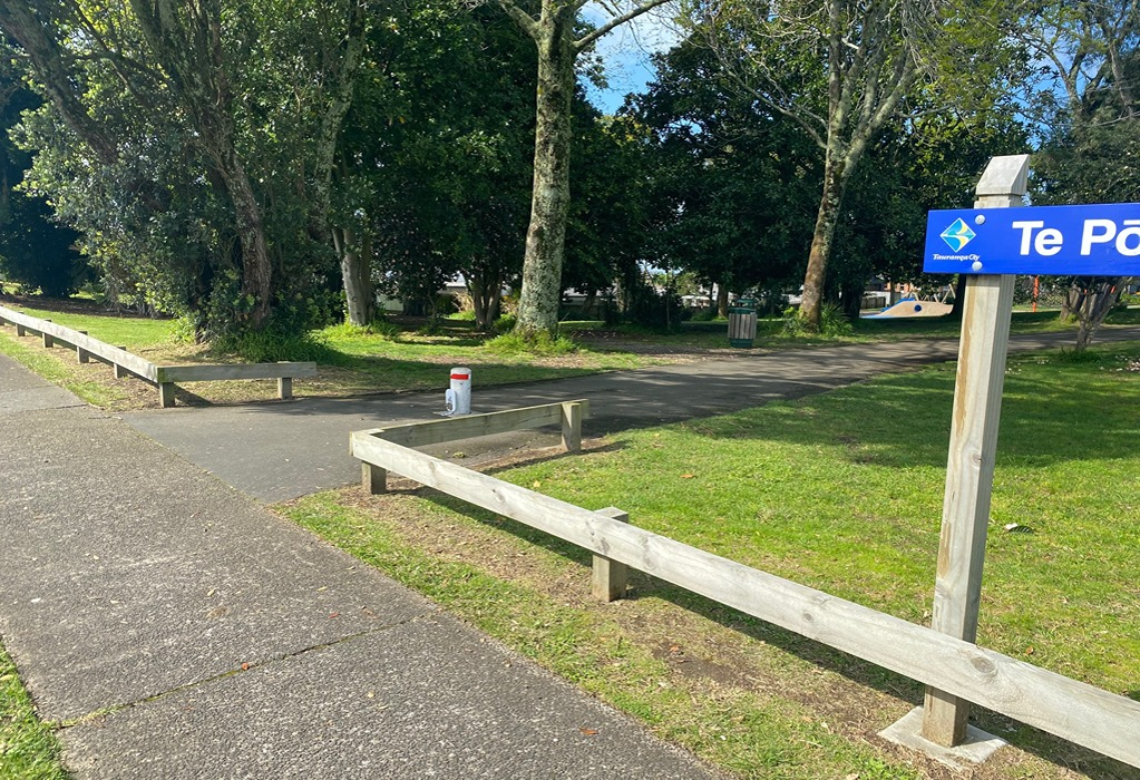 Level grey concrete and asphalt pathways with low timber fencing. A blue signpost is on the right. A white bollard with a red band is in the path centre. Grassy areas with mature trees surround.