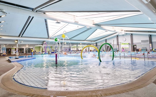 Aquatic centre with ramped beach entry pool and non-slip tiled surround. Bright natural light. Green and yellow spray hoops and silver metal handrails lead into the lap pool section.