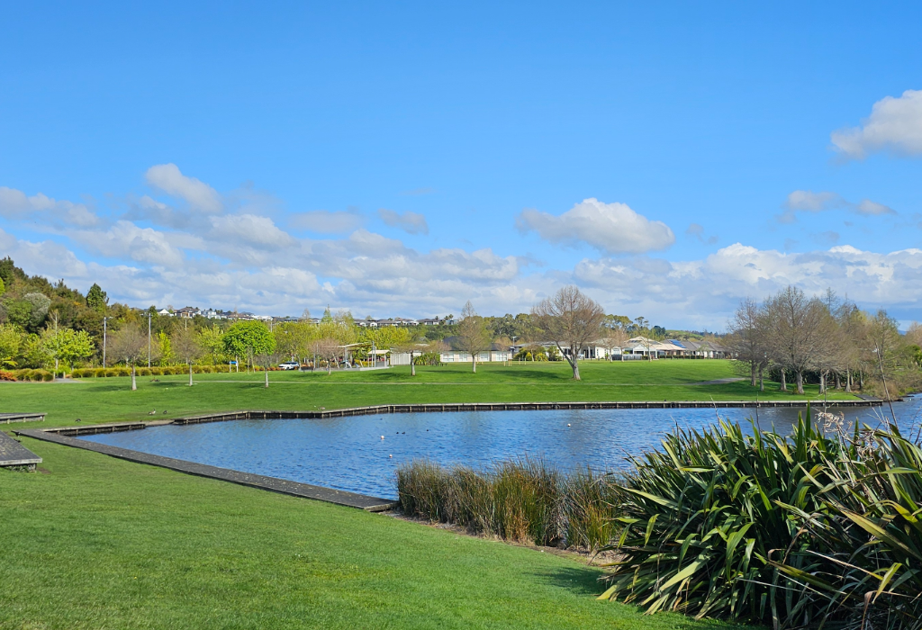 Level dark grey boardwalk edges a blue lake. A wide green grass lawn, distant paved pathway, and scattered trees provide flat terrain under a clear blue sky.