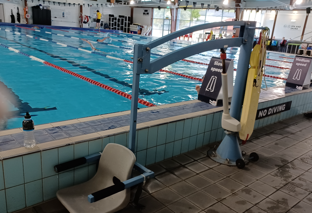 Blue pool hoist with a beige plastic seat and black armrests at the edge of a tiled indoor pool. A 'NO DIVING' sign is on the wall. Swimmers use lanes in the background.