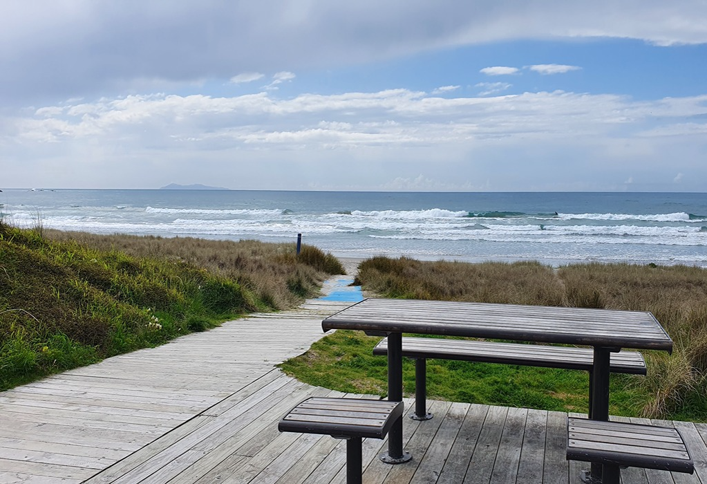 Wide, level timber boardwalk leads to a blue beach access mat. A timber picnic table with bench seats sits on an adjoining wooden deck beside coastal vegetation, overlooking the ocean.