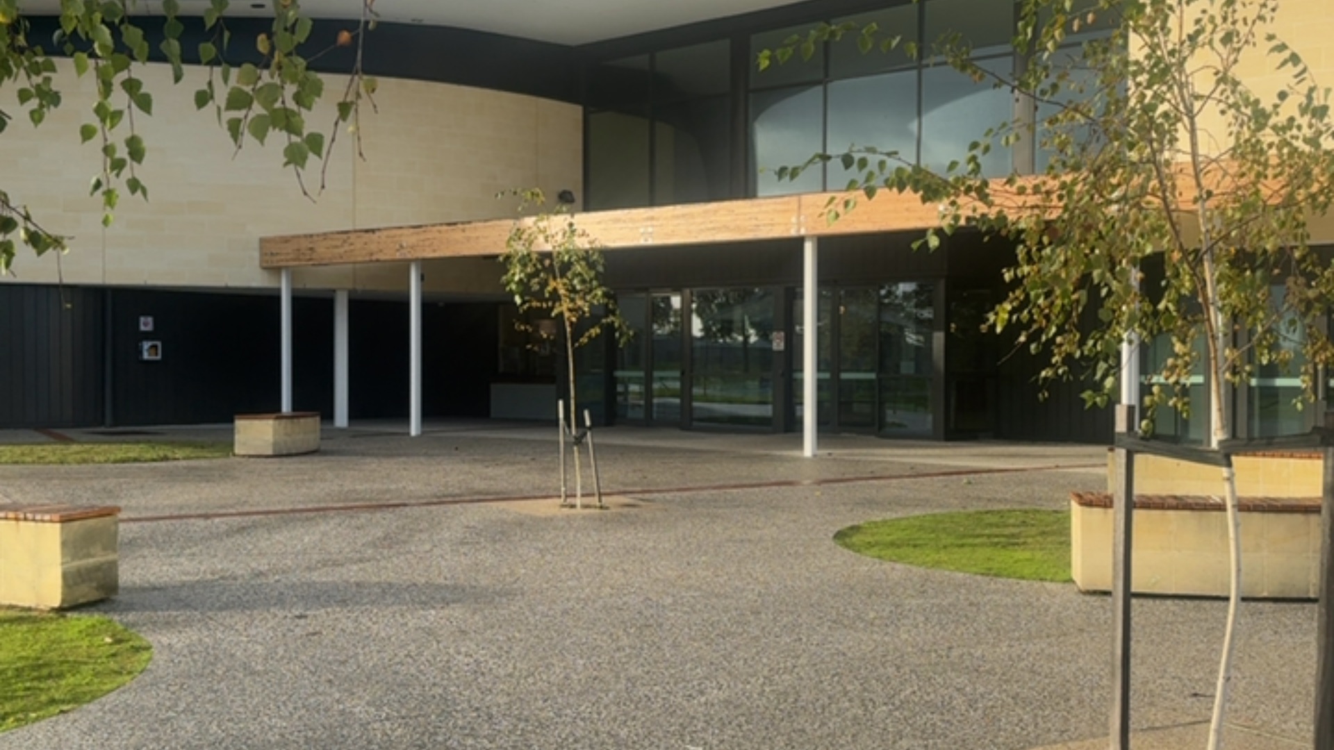 Level entrance to glass doors under a wide timber awning. Flat, textured grey concrete aggregate ground surface with low sandstone block seating topped with timber.