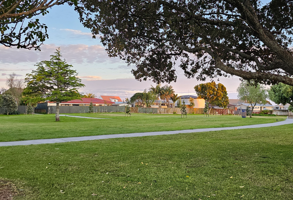 Wide, level concrete pathway curves through an unobstructed, flat grassy park. Mature trees and houses line the perimeter. A blue bin is located beside the path. Natural evening light.