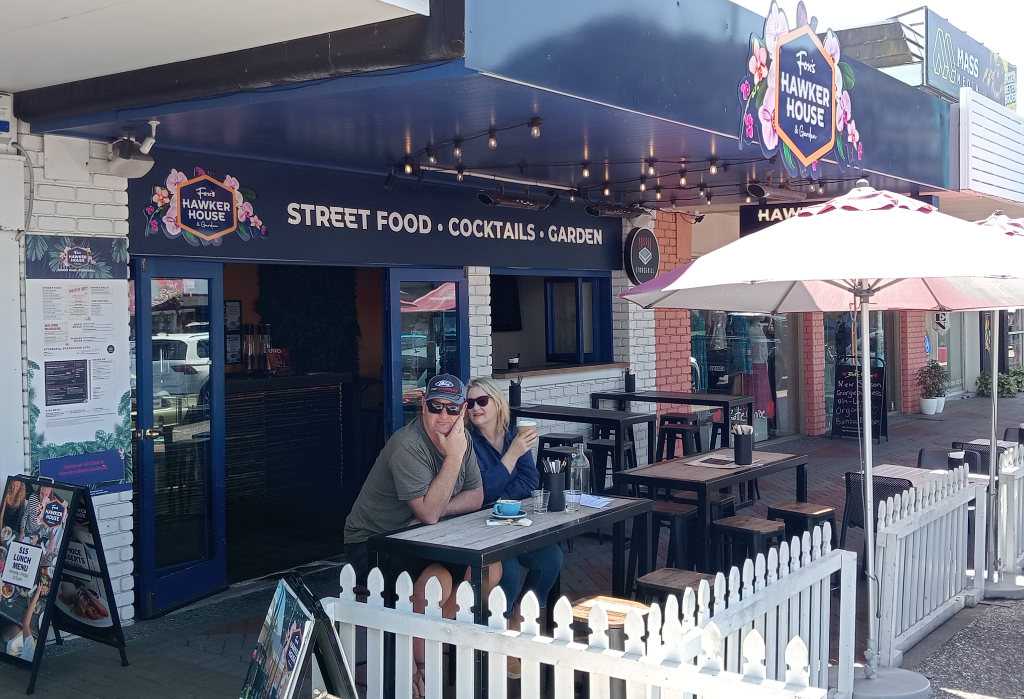 Level paved outdoor dining with high-top timber tables and stools behind a white picket fence. Glazed entrance has a small threshold. A-frame signs sit on the path under a blue awning.
