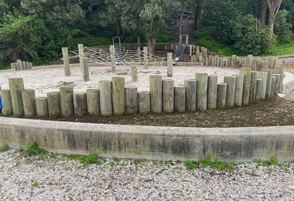 White sand play area enclosed by a curved concrete wall and timber logs. Features a rope climbing net and metal slide. Level gravel ground in foreground meets the wall; no ramp is visible here.