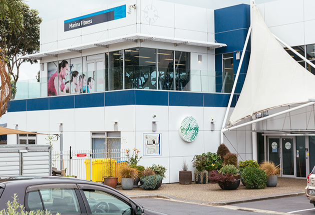 Step-free glass double doors under a white sail awning. Level bitumen surface leads to the entrance of a two-storey white and blue building with signage for Marina Fitness and Saplings.
