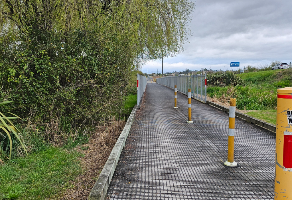 Level, wide walkway with a grey metal mesh surface and metal fencing on both sides. Yellow bollards with white reflective bands are spaced along the right side of the path.