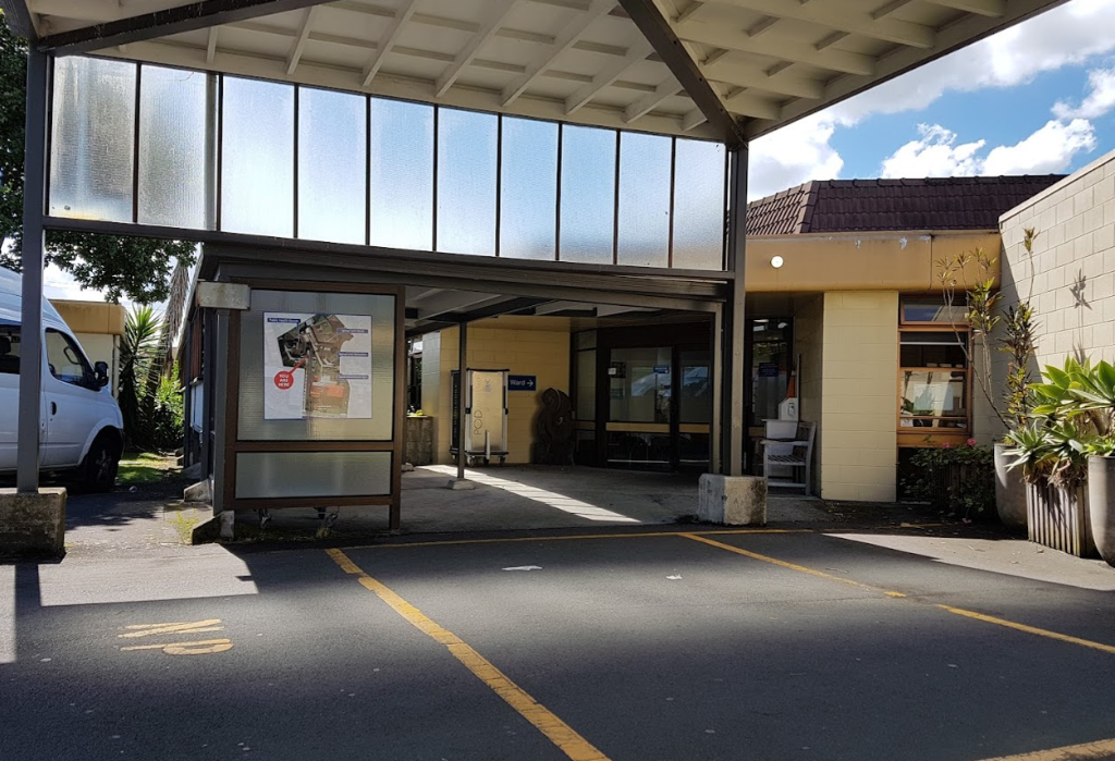 Level asphalt ground with yellow markings leads under a covered walkway to a glass-door entrance. A directory map is on the left and a blue sign points to the Ward on the right.