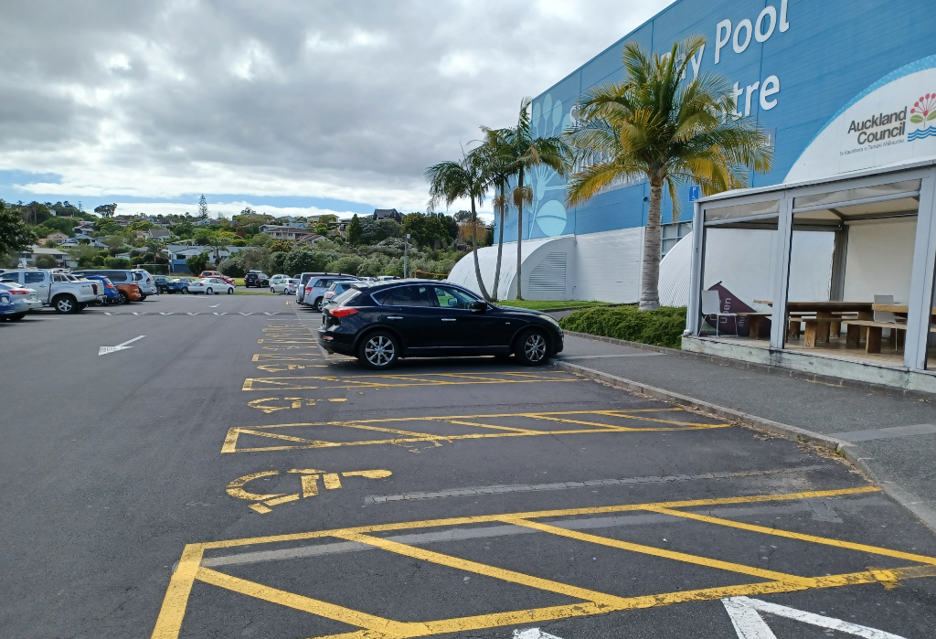Level bitumen car park with accessible spaces marked by yellow symbols. A black car occupies one. A concrete kerb and pathway lead to a blue building with a covered outdoor seating area.