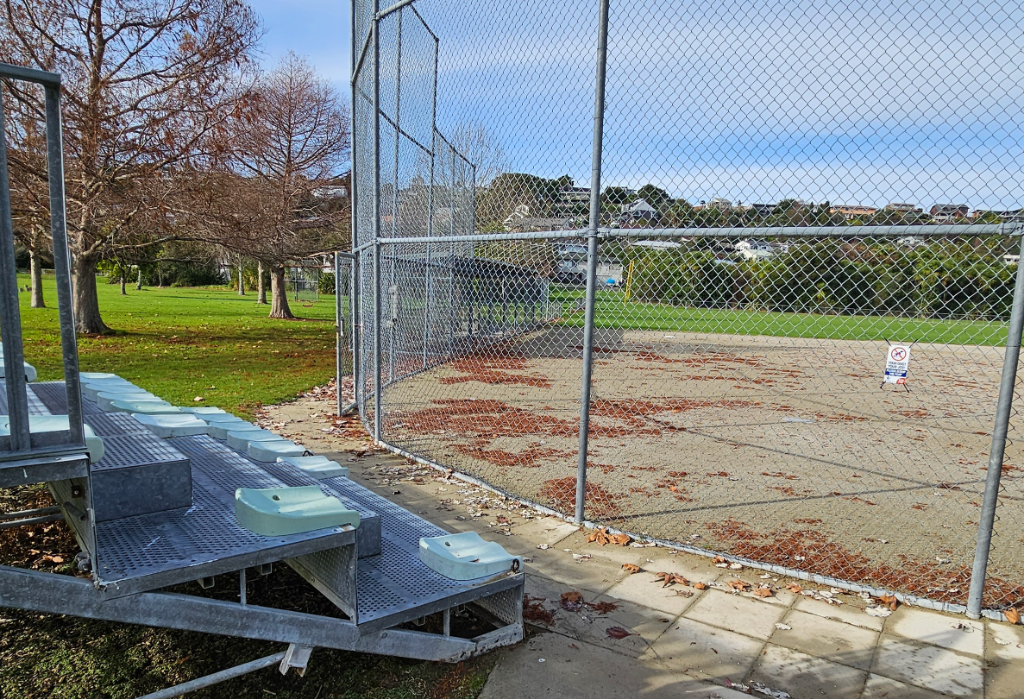 Metal grandstand with tiered green seats and galvanised steel steps beside a level paved path. A high chain-link fence encloses a dirt sports field scattered with fallen leaves.