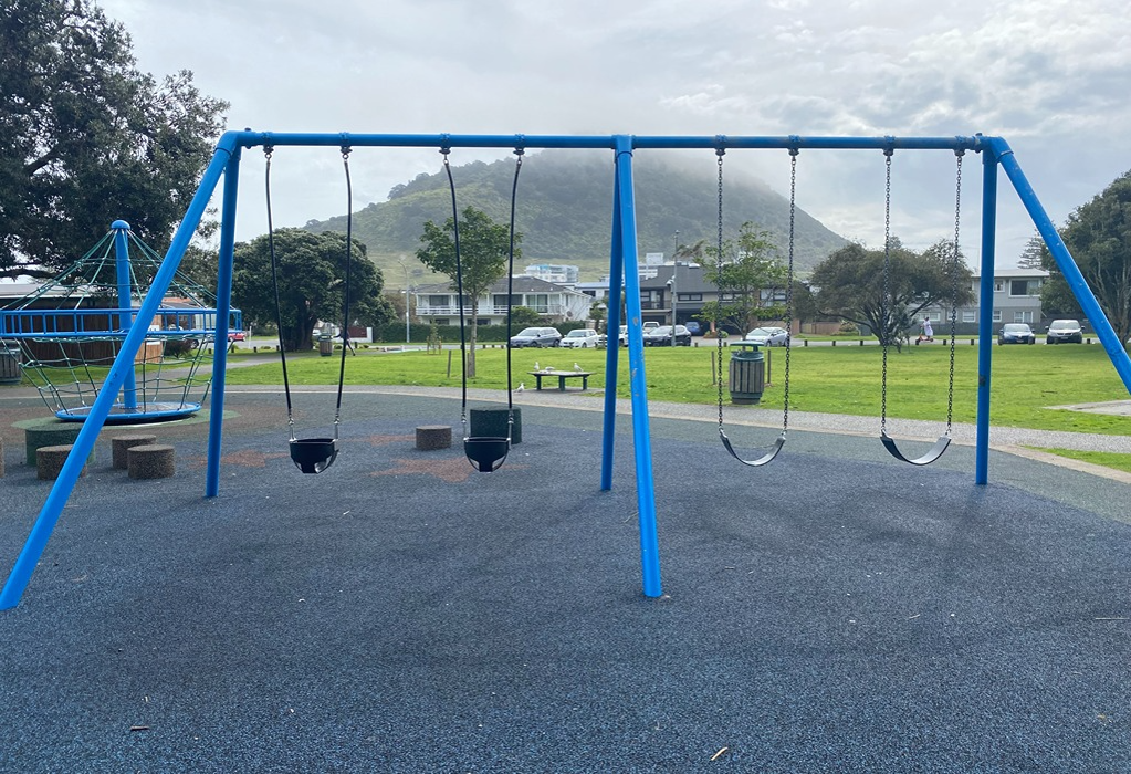 Blue swing set and rope climbing frame on level dark grey rubberised safety surface. Level concrete pathway borders green grass. Concrete seating blocks nearby. Misty hill in the background.
