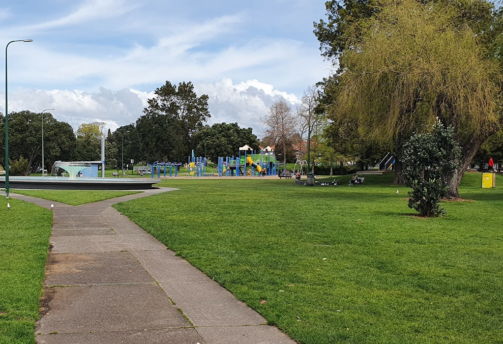 Wide, level concrete pathway forks across flat grass toward a playground with blue and yellow equipment. A circular fountain and park benches are situated under mature trees.