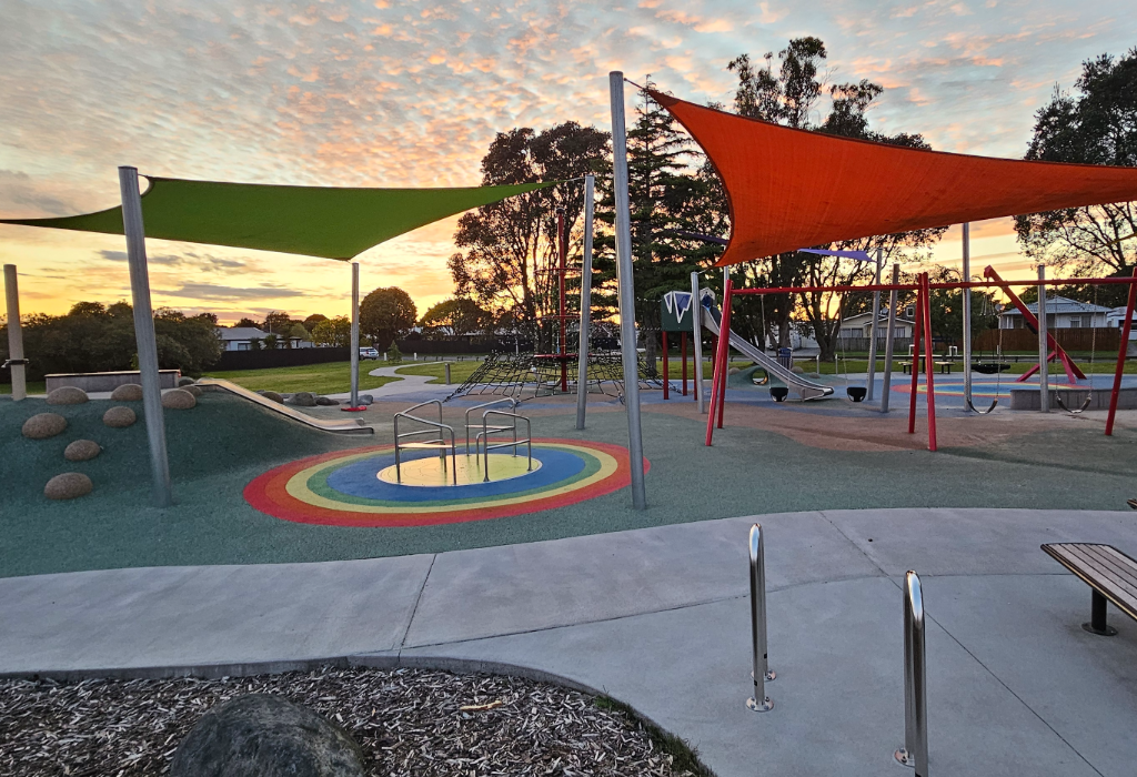 Level concrete path leads to a playground with colourful soft-fall rubber flooring. Includes a wooden bench, metal handrails, and play equipment under green and orange shade sails.