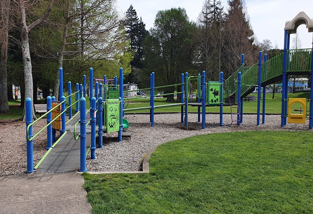 Level concrete path joins a metal mesh ramp with green handrails and blue poles, accessing a raised playground structure. Ground surfaces include grey gravel and green grass.