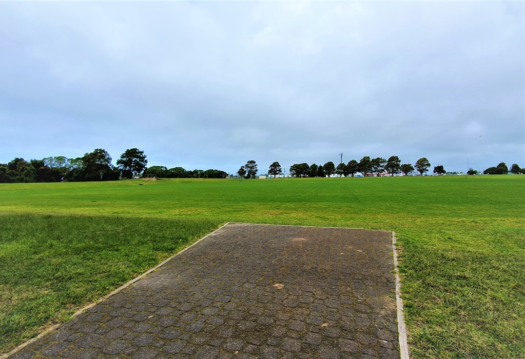 Level, wide rectangular pad of dark grey textured pavers with light-coloured edging, set flush with a large, flat, open grass field bordered by distant trees.