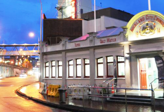 Entrance with stairs and a ramp with silver handrails. Wet concrete footpath with yellow safety barriers. Heritage building facade with lit windows and 'Tepid Salt Water' signage.