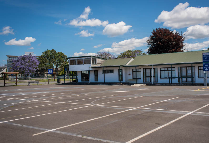 Level asphalt area with white markings leads to a building with a small concrete ramp to its porch. A blue 'Please Pay Here' sign stands near a ticket machine on the right.