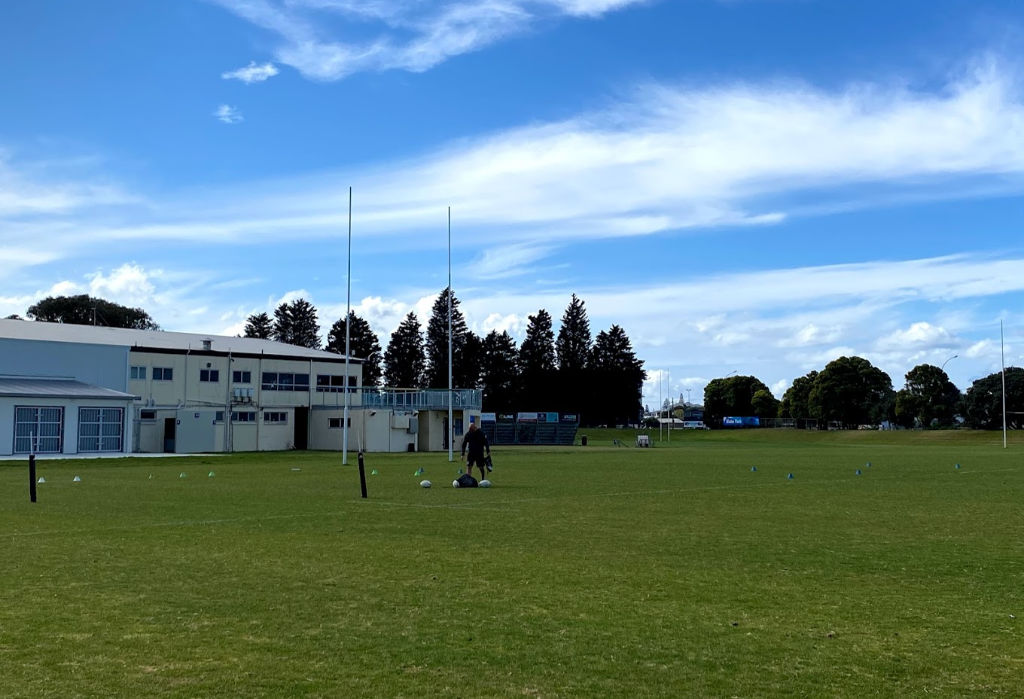 Level green grass sports field with tall white goal posts. A light-coloured building features a level concrete apron. Small tiered grandstand and blue signage in the distance.