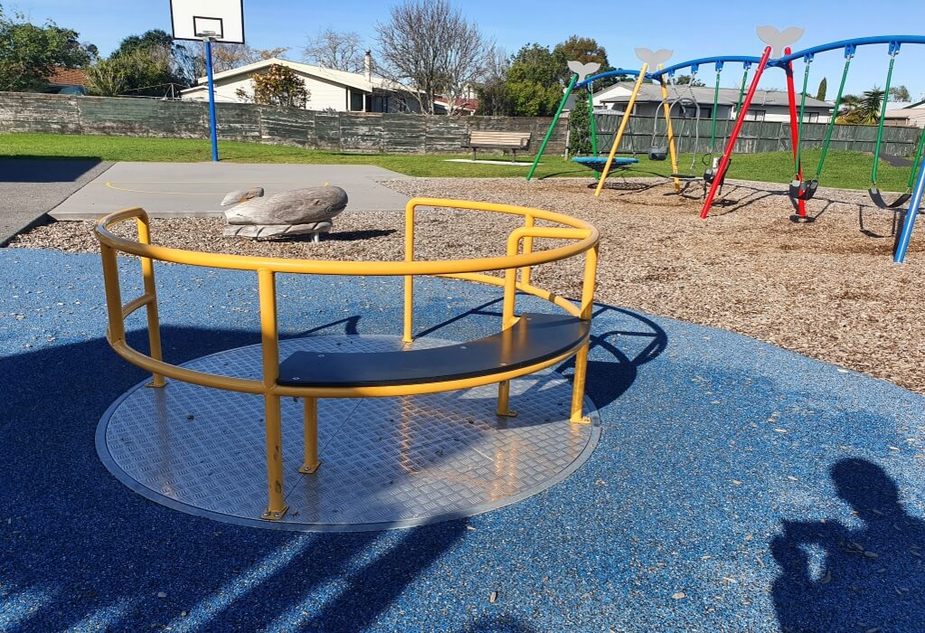 Yellow merry-go-round with black bench, level with blue rubber safety surfacing. Wood chip area behind with multi-coloured swing set, concrete basketball court, and wooden park bench.